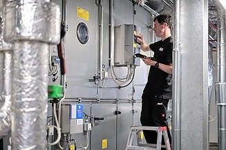 Photo of an employee on a ladder at the ventilation system, transferring data to a tablet 