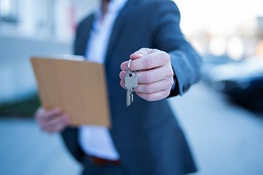 Man in blue suit and white shirt, clipboard in hand, hands over bunch of keys. 