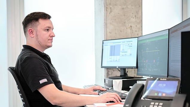 Man in a shirt at a desk with three screens, telephone with display on the right edge of the picture.