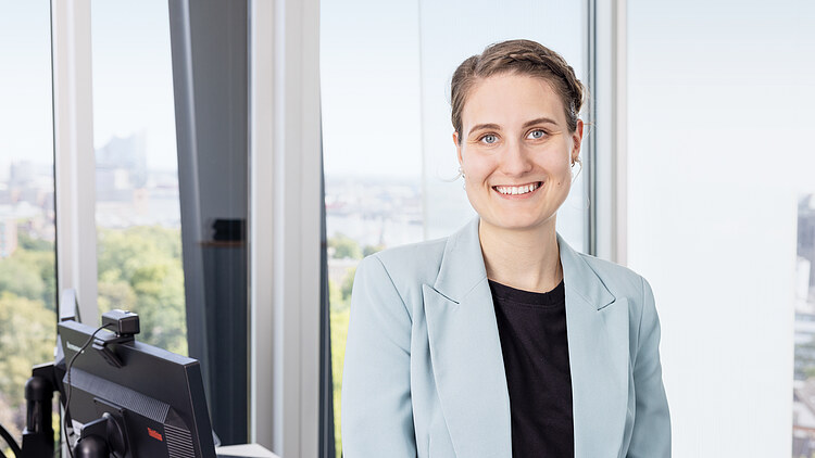 Portrait von Mona Gärtner - Freundliche Frau im hellblauen Blazer lächelt in modernem Büro mit Ausblick auf Stadtlandschaft, Business-Porträt, professionell.