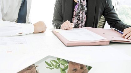 Zwei Personen prüfen Verträge am Schreibtisch in einem Büro, Nahaufnahme von Dokumenten, Händen und einem Tablet.