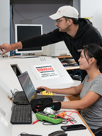 Foto von zwei Mitarbeitenden an einem Tisch mit Monitoren und einem Laptoptop-