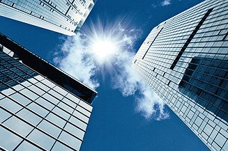 Hochhäuser aus Glas ragen in den blauen Himmel, die Sonne scheint zwischen den Gebäuden hindurch.