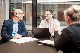 Drei Kollegen im Meeting, zwei Männer im Gespräch mit Laptop, Frau von hinten, moderne Büroumgebung.